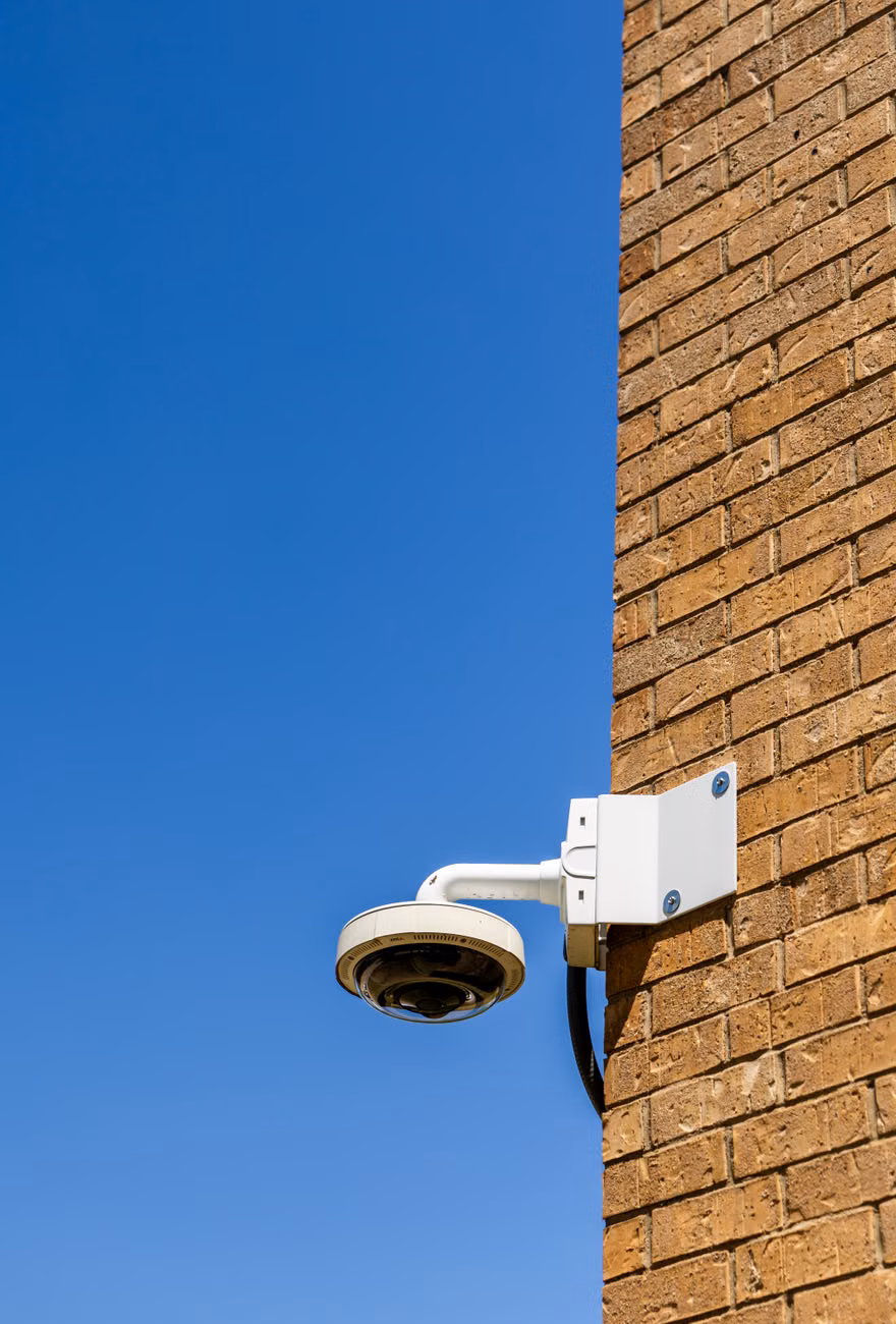 A security camera mounted on a brick wall against a bright blue sky, capturing urban surveillance.